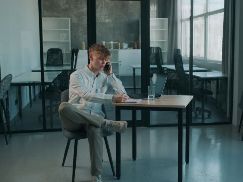 momak sedi za stolom u kancelariji i priča na telefon i unosi podatke u notes