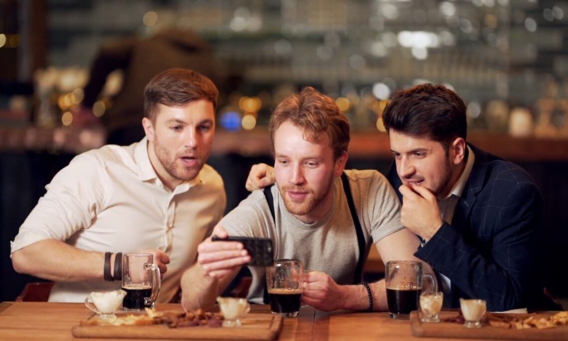 tri muškarca sede u kafiću, piju crno pivo i gledaju sportsku utakmicu preko telefona