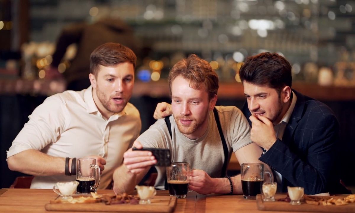 tri muškarca sede u kafiću, piju crno pivo i gledaju sportsku utakmicu preko telefona
