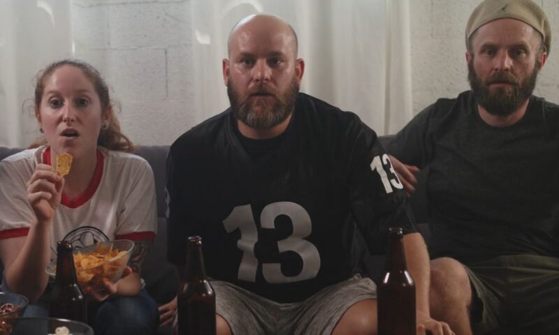 dva momka i devojka sede na ugaonoj, piju pivo, grickaju grickalice i gledaju uživo utakmicu